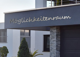 Dies ist ein Bild mit einem Schriftzug aus Edelstahl MÖGLICHKEITENRAUM aus der Kategorie Firmenschilder an moderner Fassade